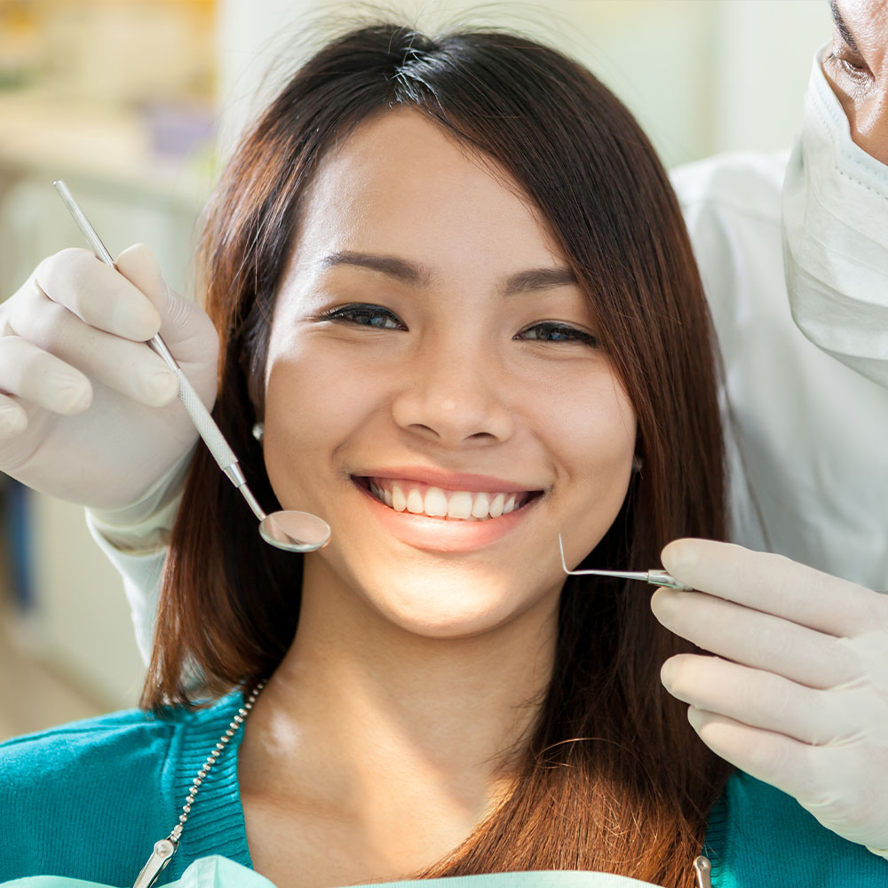 A woman sitting in a dental chair with a smiling expression while receiving dental care, assisted by a dental professional wearing gloves and a mask.