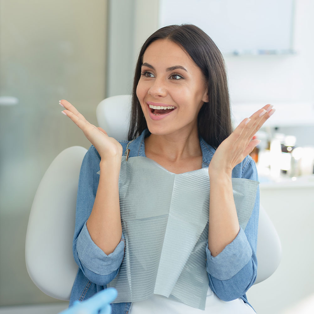 A woman with long dark hair wearing blue jeans, standing in front of a dental chair, smiling broadly with her mouth open wide, posing for a photograph.