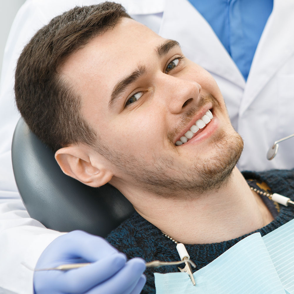 A young man with a beard is sitting in a dental chair, smiling broadly, with his teeth being worked on by a dental professional who is wearing gloves and a mask.