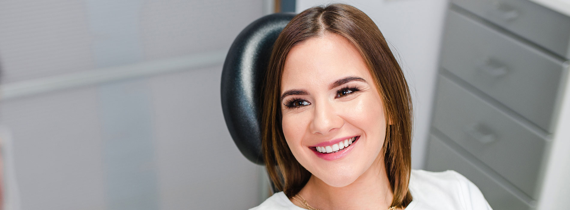 Woman sitting with hands clasped together, smiling at camera, wearing white shirt, in front of blurred office setting.