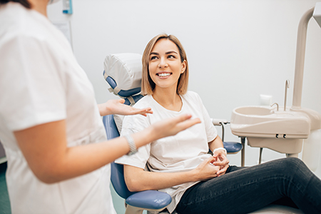 A woman with short hair sits in a dental chair, smiling at a female dentist who stands beside her.